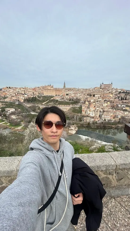 Tomokichi standing on a hillside while travelling, with a river and an old town on the ridge behind